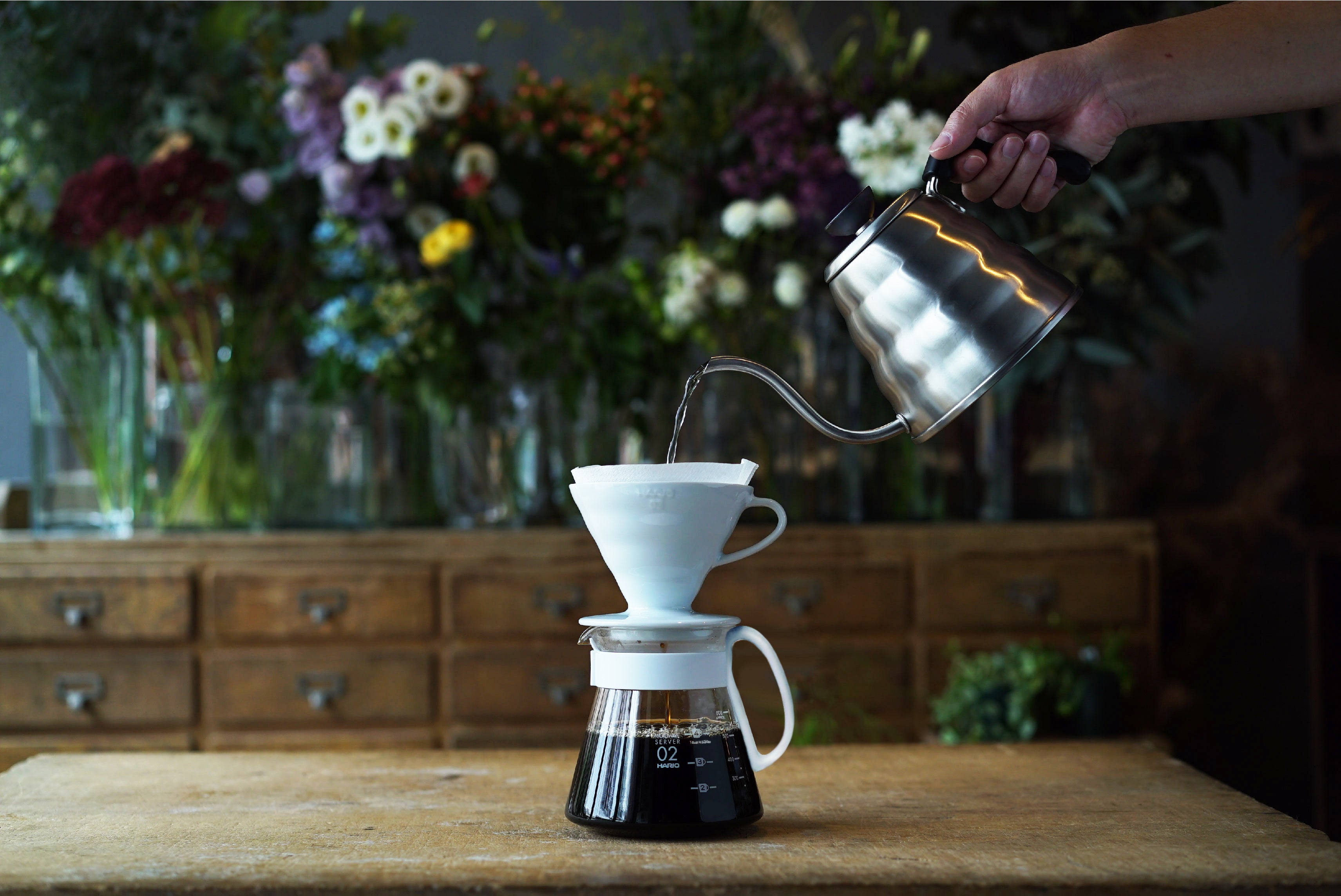 A photo of a person pouring water from a silver long spout kettle into a V60 filter and decaf coffee dripping into the jug below.