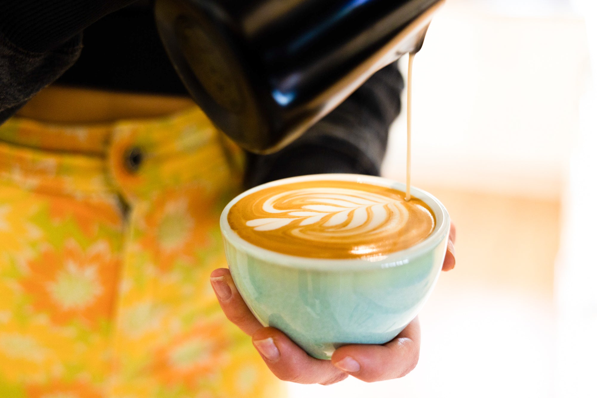 A cup with a beautiful rosetta latte art being completed, in the hand of a woman wearing bright trousers.