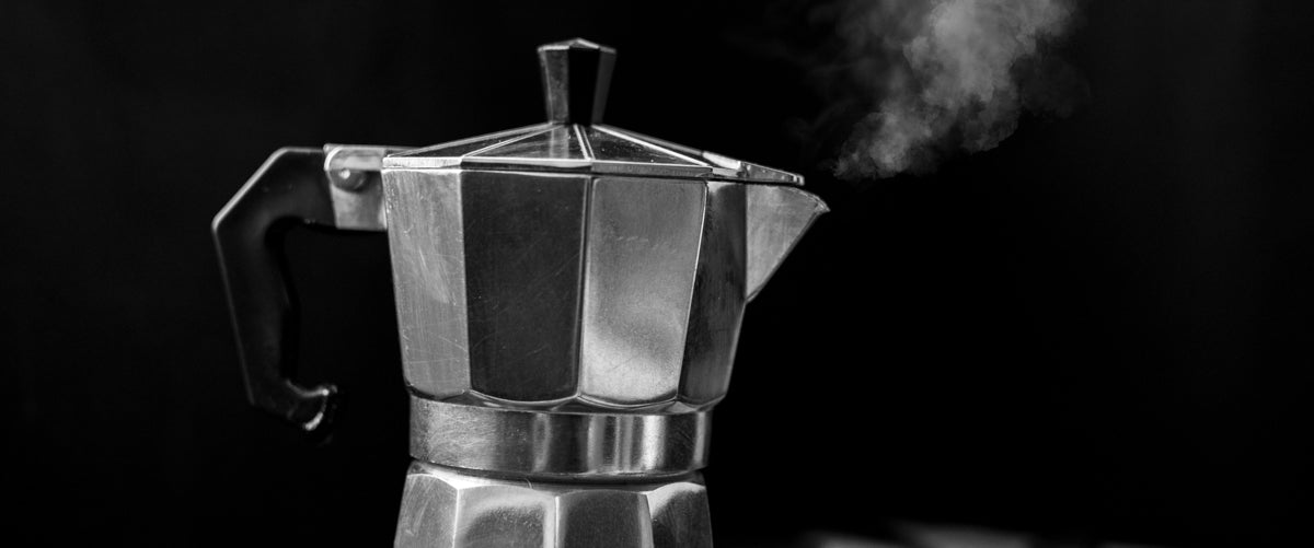 A silver moka pot with steam coming out of the spout with a black background