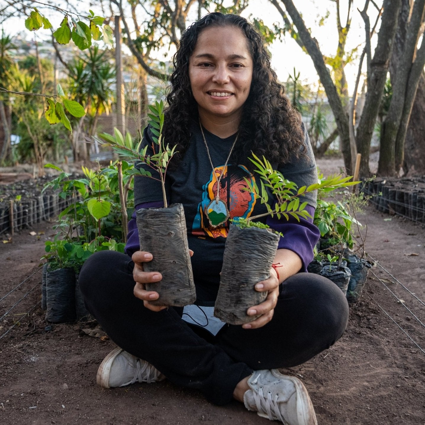 A lady with dark curly hair sat cross legged in a farm holding two small plants.