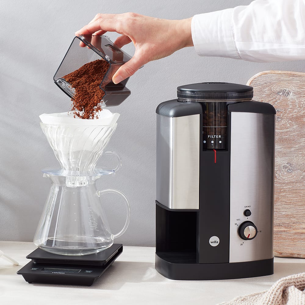 The Wilfa svart coffee grinder with the ground coffee being poured into a filter coffee setup by a hand.