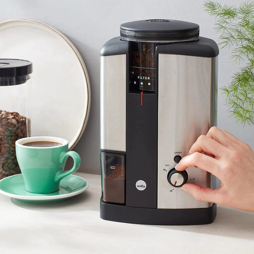 The silver wilfa svart coffee grinder being used by a hand, next to a green cup of coffee.