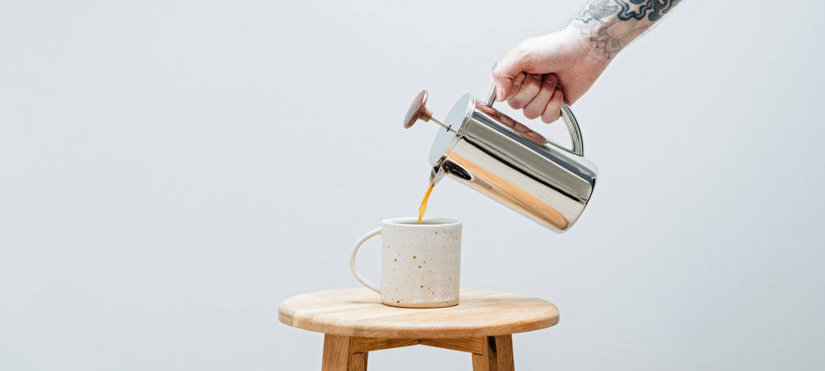 A chrome cafetiere pouring decaf coffee into a mug in a stool, being poured by a tattooed arm.