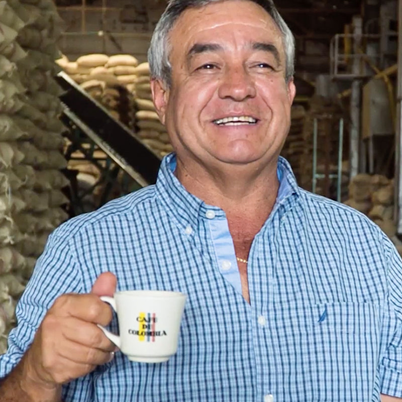 A picture of a man wearing a blue checked shirt, smiling, holding a cup.